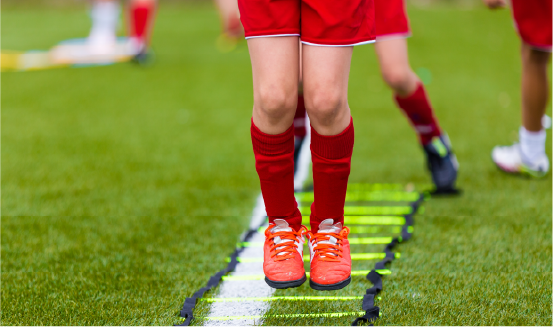 youth-athlete-agility-ladder-training-red-uniform Close-up of young athlete's legs in red soccer uniform and cleats performing agility ladder training drill on grass field, showing proper youth sports conditioning technique